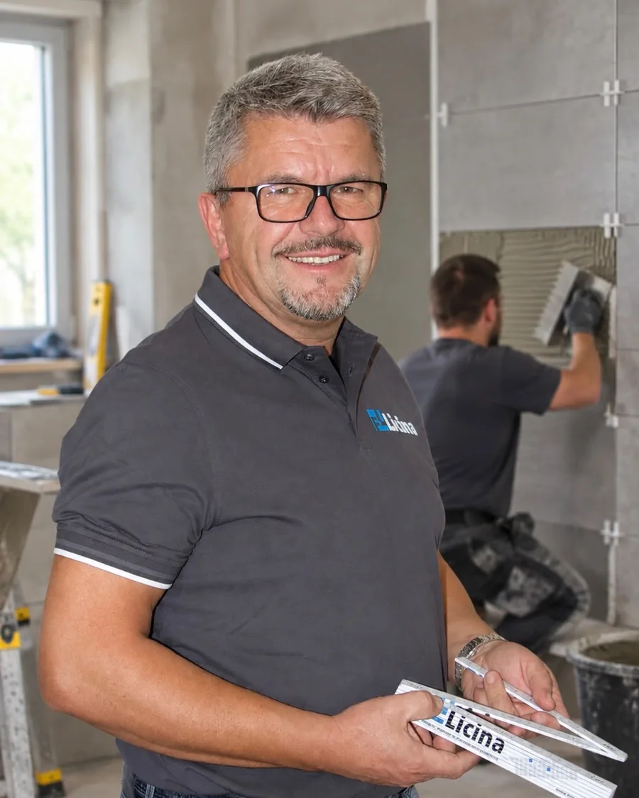 Fahrudin Licina, Inhaber von Fliesen-Licina, mit Zollstock auf einer Baustelle, im Hintergrund ein Mitarbeiter beim Fliesenlegen